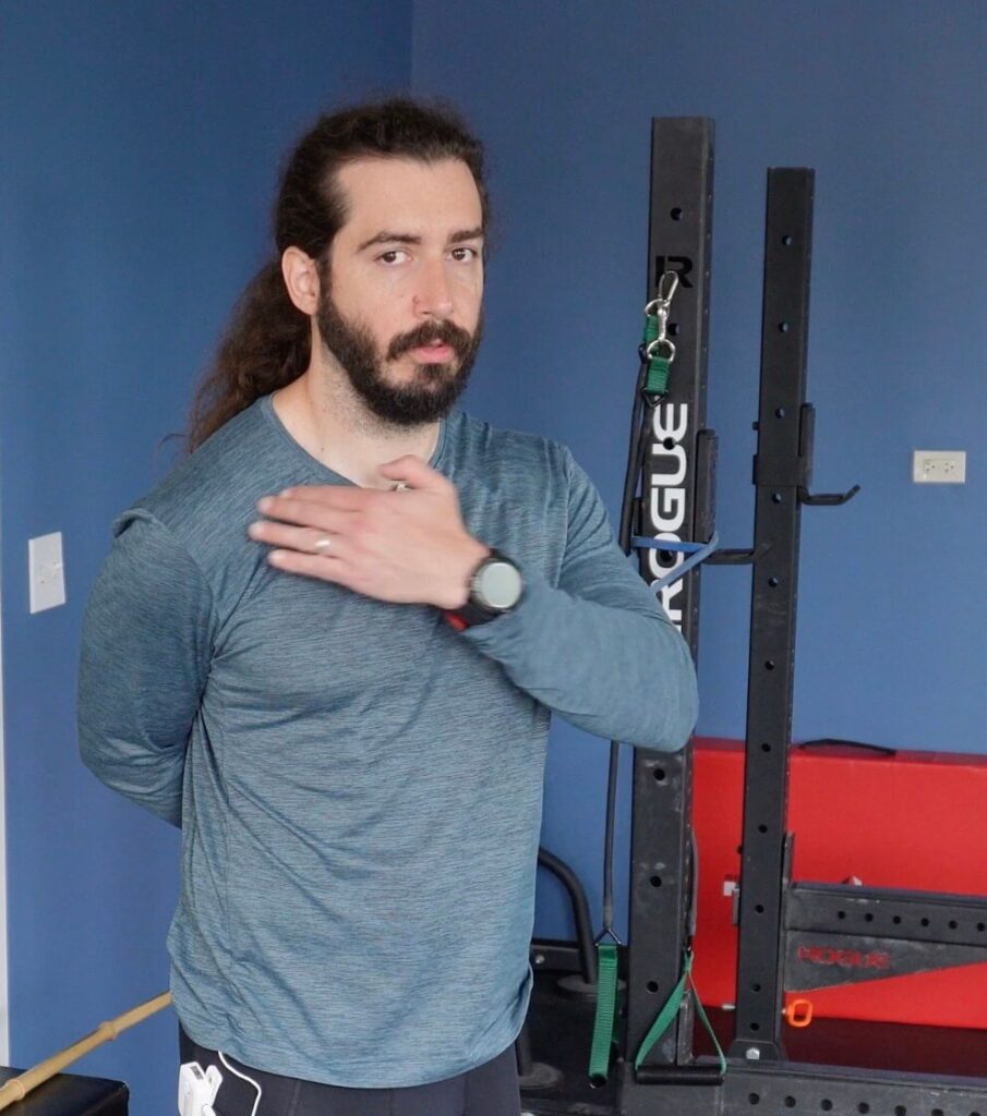 Dr. John with his right hand being pulled behind his back by a band attached to a squat rack. He is improving his internal rotation for his front rack position